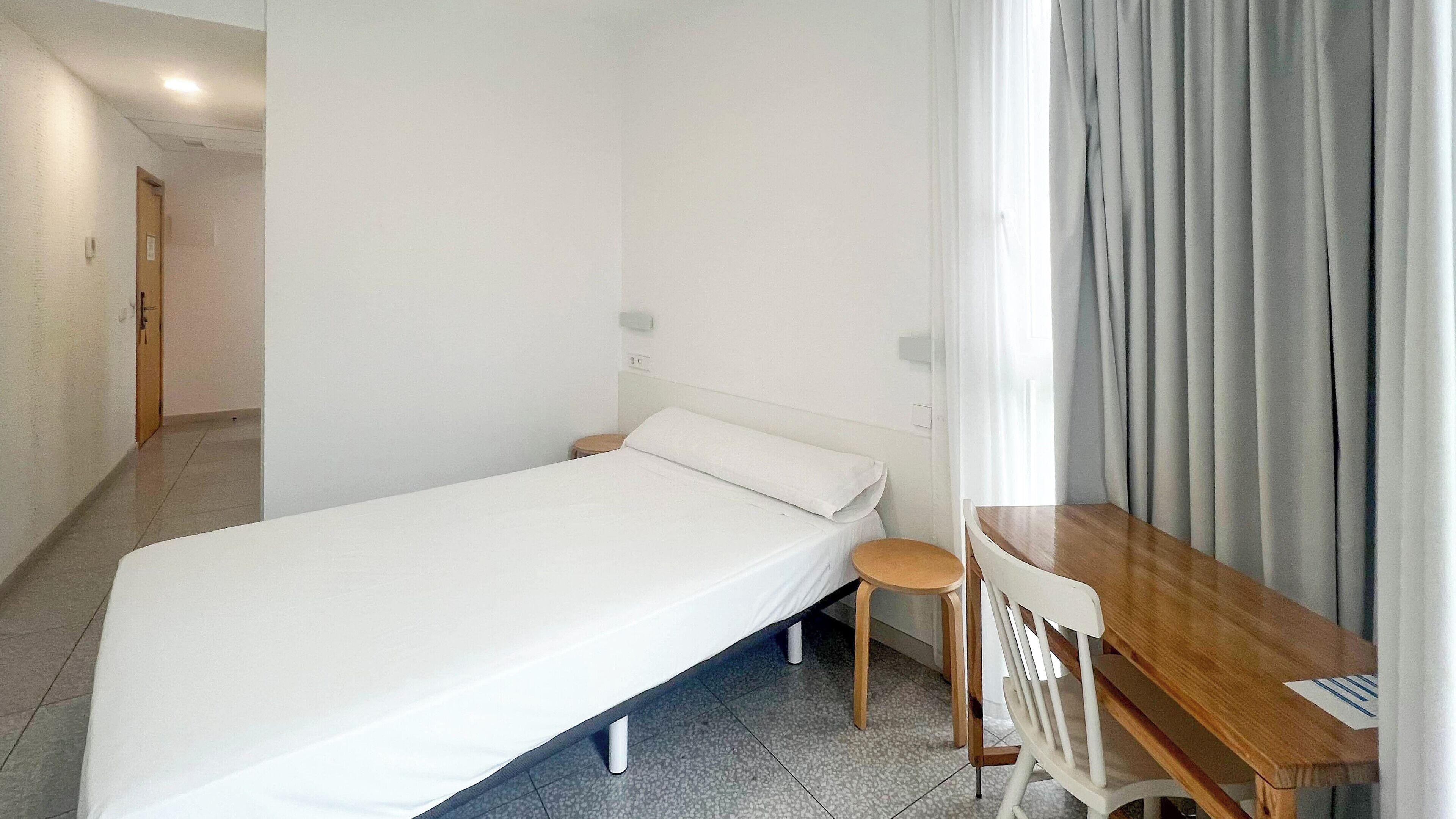 A small, clean room with a single bed, a wooden table, and a chair.