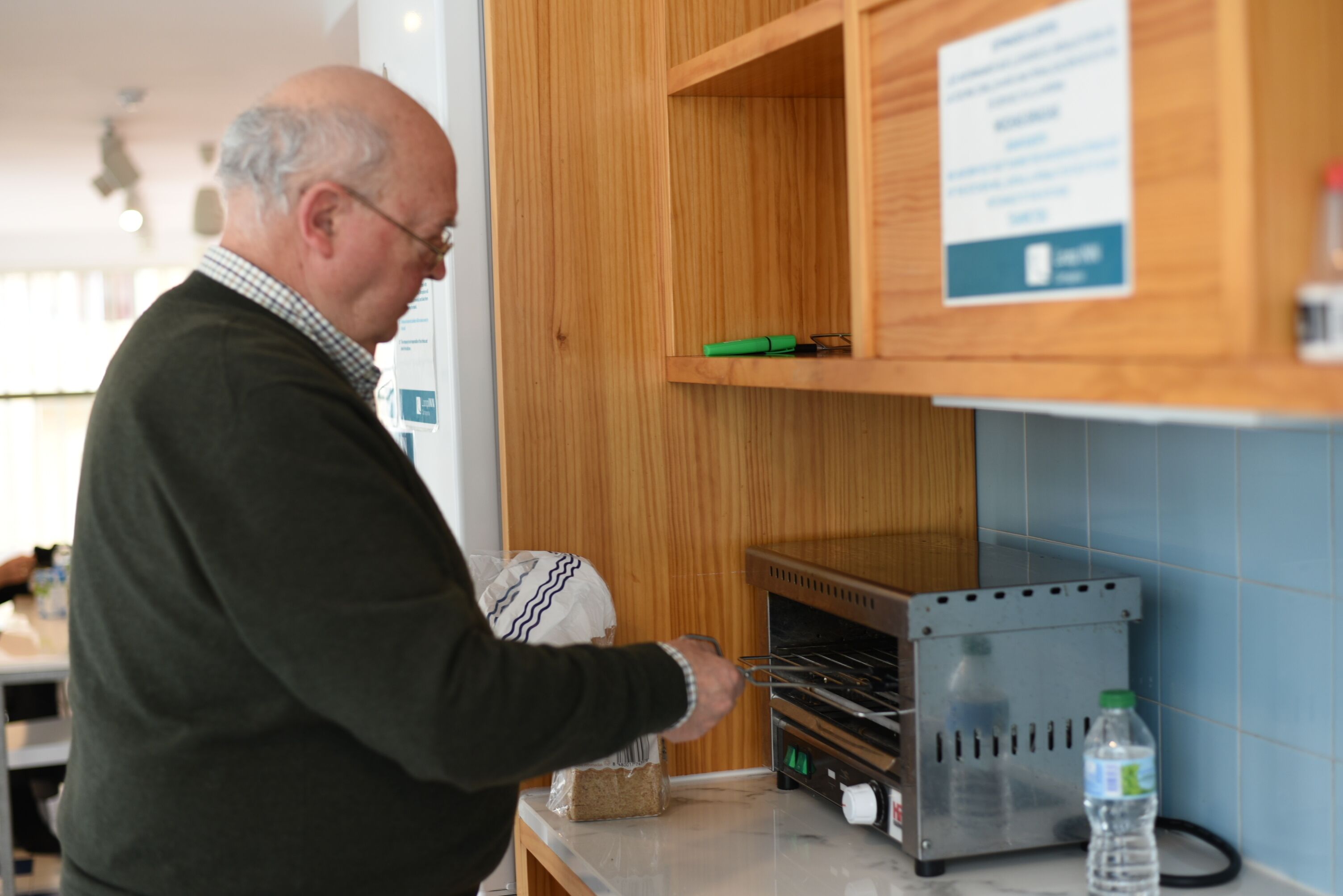 A man in a kitchen with a bread box, a bread box on the counter, and a bottle of water.