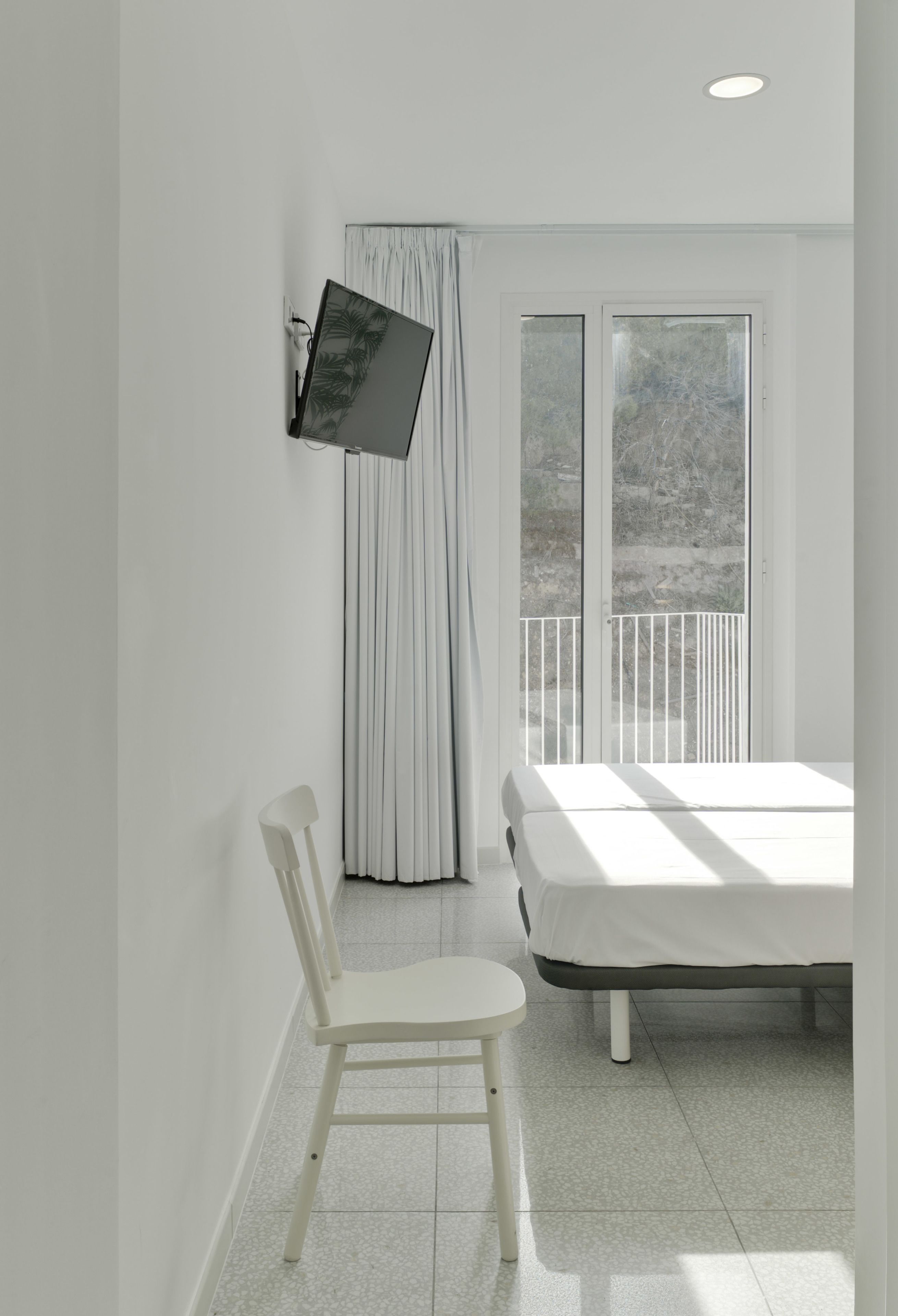 A bedroom with a bed, a chair, a TV mounted on the wall, and a balcony with curtains.