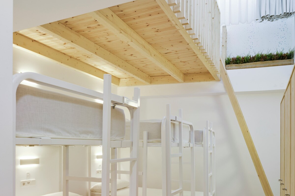 A children's bunk room with white bunk beds, a wooden staircase, and a small window with curtains.