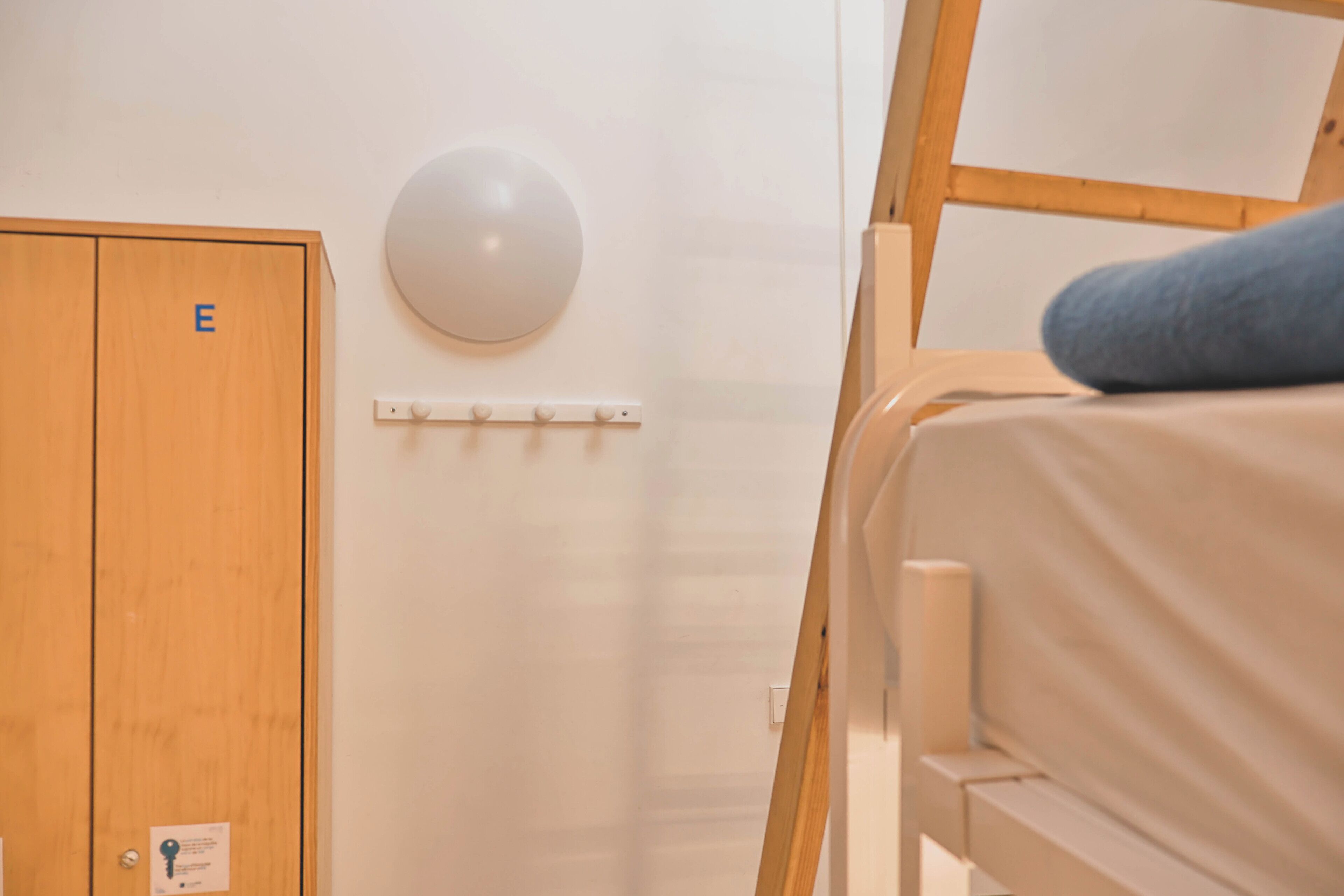 Una cama litera con un cojín, un armario de madera con etiqueta 'E', una lámpara de pared y una pared blanca.