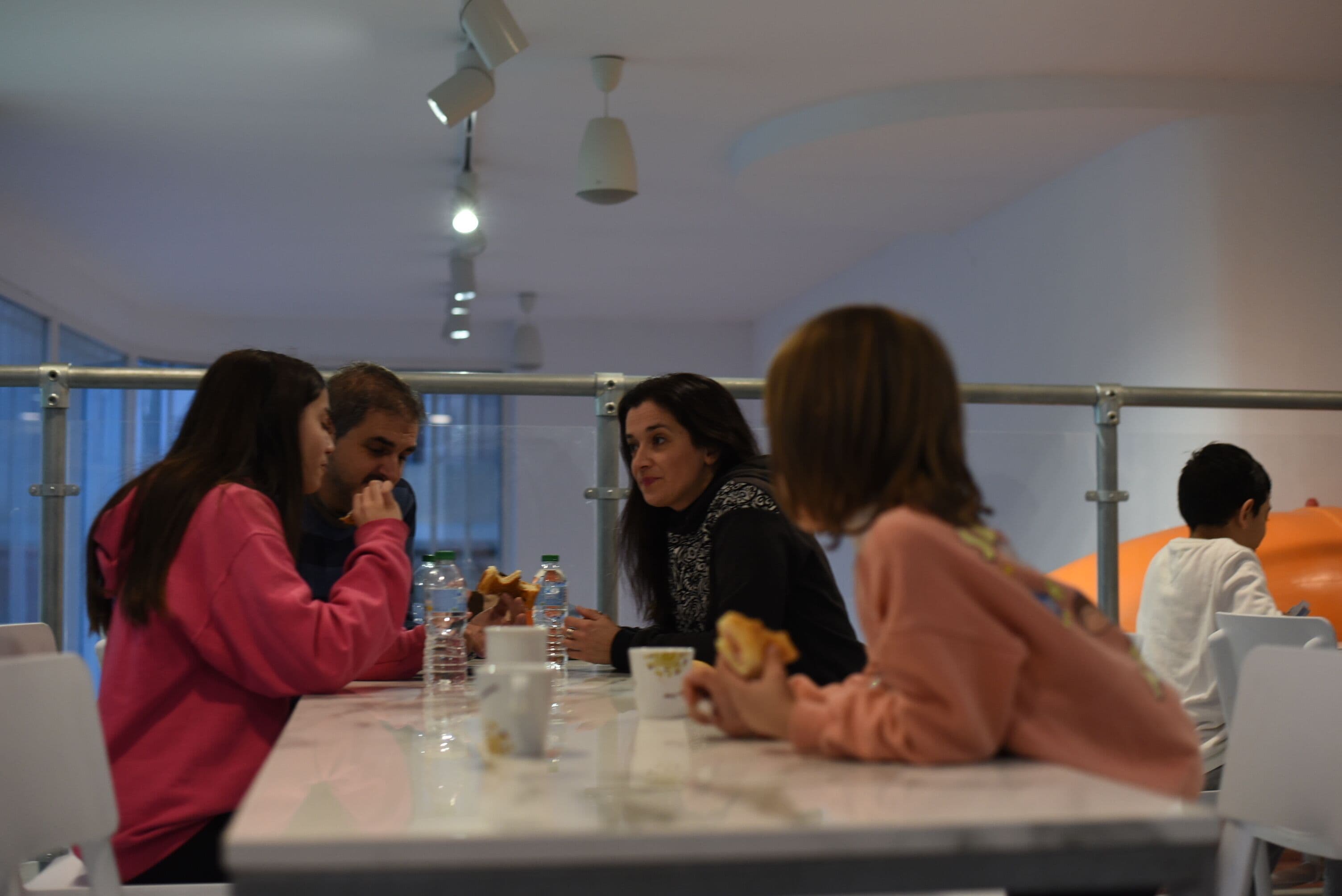 A group of people seated at a table in a modern dining area with a high ceiling and pendant lighting.