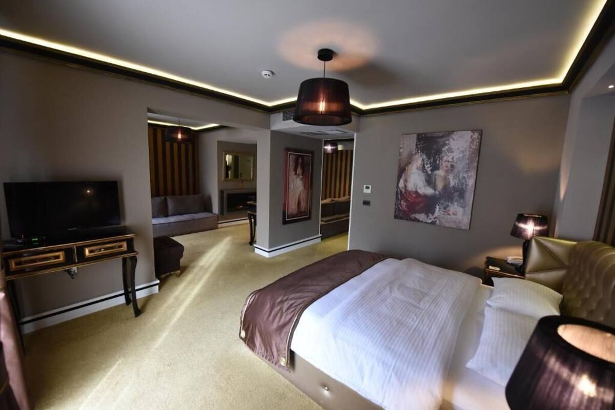 A modern hotel room with a large bed, a flat-screen TV, a desk, and a seating area with a chair and a lamp.