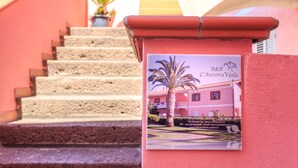 A property with a staircase leading to a pink building, a signboard with contact information, and a potted plant. -  L'Aurora Viola (Carbonia)
