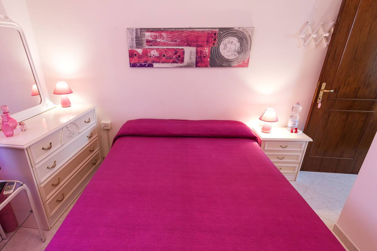 A bedroom with a pink bedspread, a dresser with drawers, a mirror, a wall art piece, a bedside table with a lamp, and a door.
