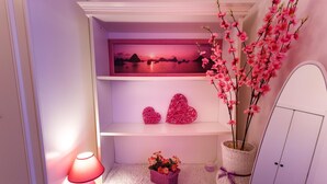 A room with a white cabinet, a wall-mounted shelf with decorative items, a small basket with flowers, and a pink lamp. -  L'Aurora Viola (Carbonia)