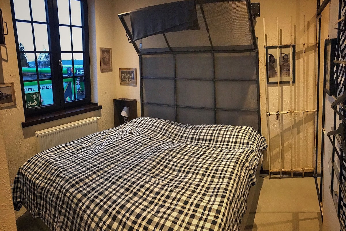 A bedroom with a checkered bedspread, a window with a view, a radiator, and a door with a metal gate.