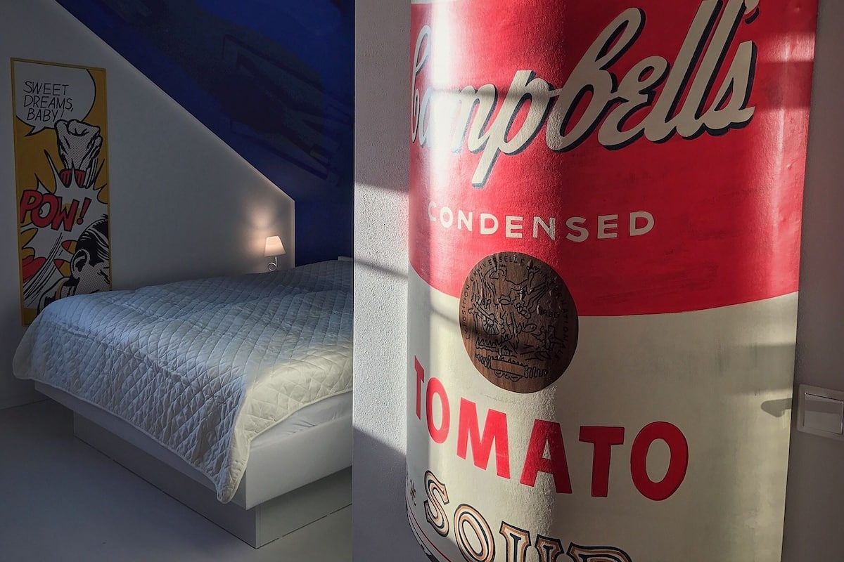 A room with a bed, a wall with a comic book-style poster, and a large can of Campbell's Tomato Soup.
