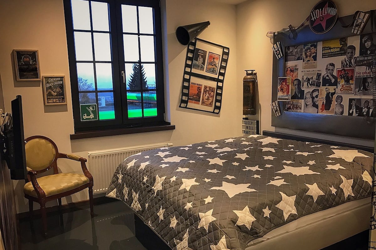 A bedroom with a bed, a chair, a window, and a wall decorated with movie posters.