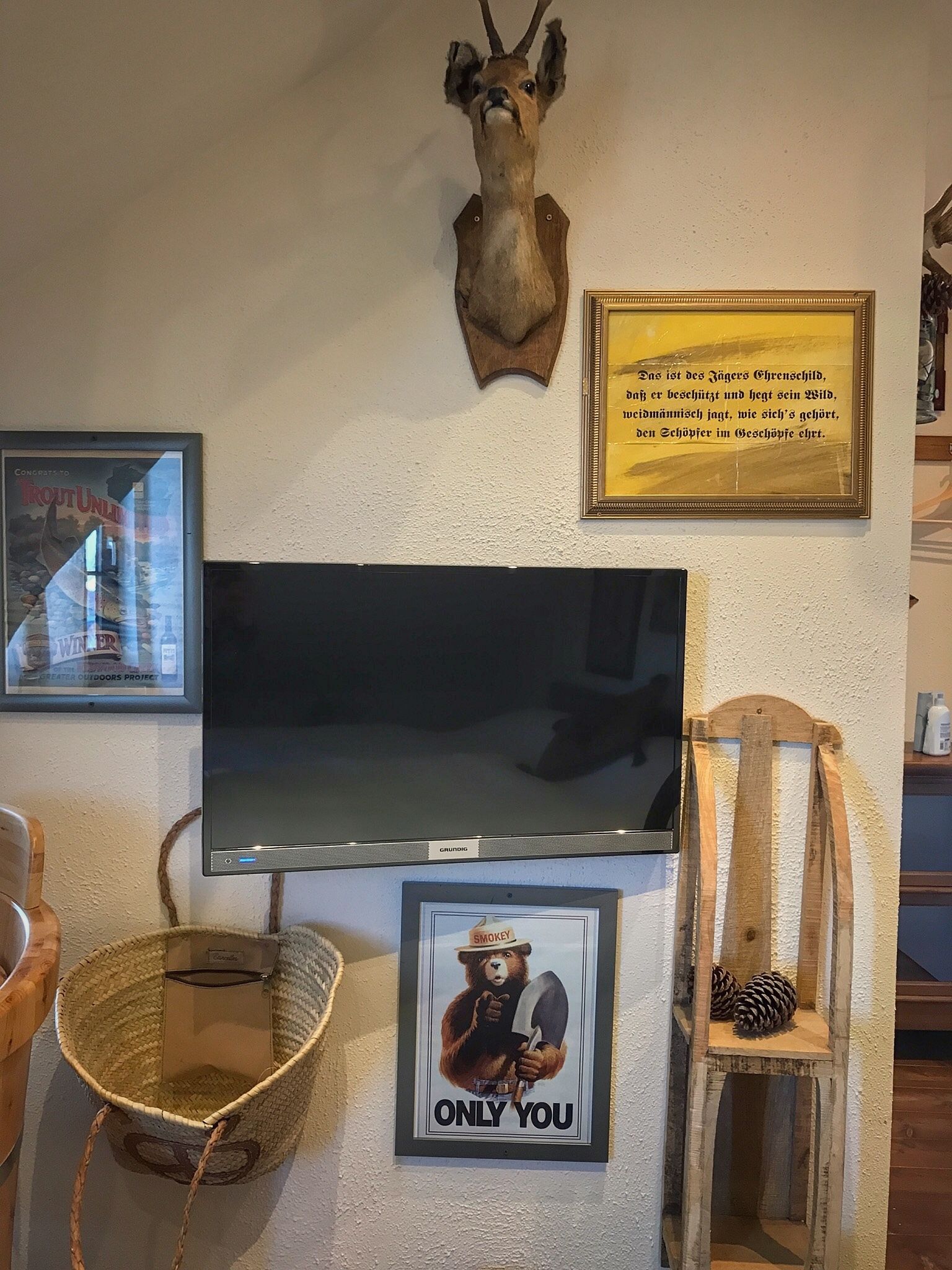 A wall with a mounted deer head, a framed quote, a framed bear picture, a television, and a woven basket.