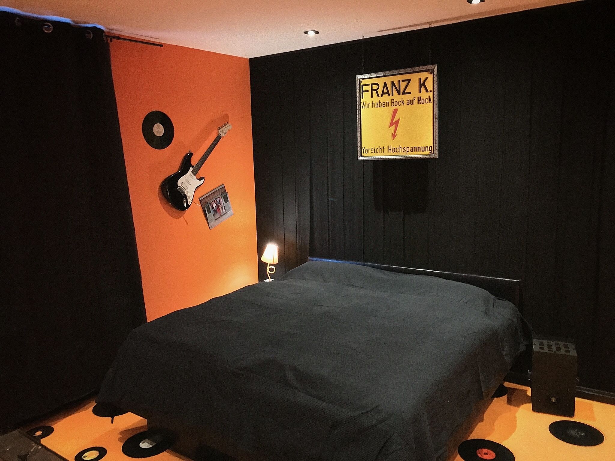 A room with an orange accent wall, a bed with a black cover, a guitar, a framed picture, and a sign with text.