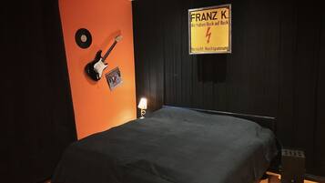A room with an orange accent wall, a bed with a black cover, a guitar, a framed picture, and a sign with text.