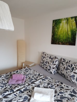 A bedroom with a bed, a nightstand, a pillow, a blanket, and a painting of a forest. - Zweite Heimat (Uebersee)