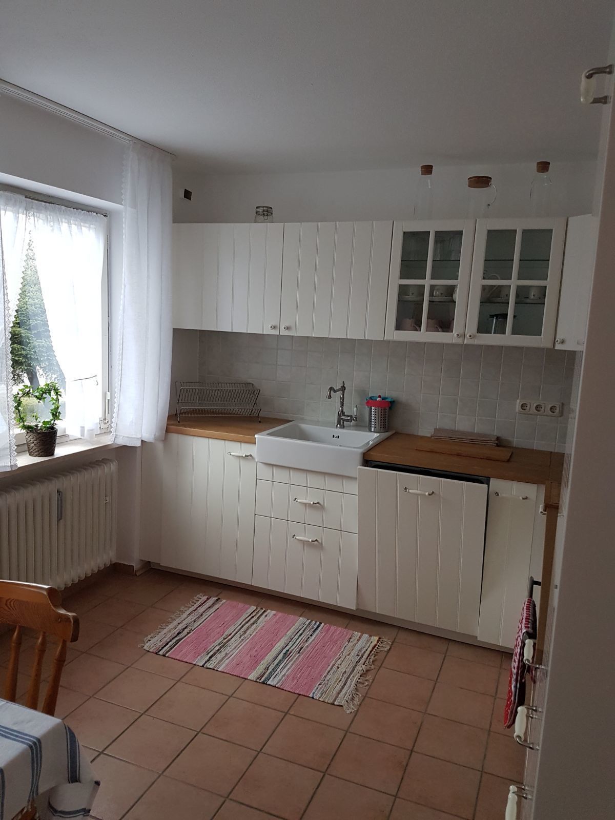 A kitchen with white cabinets, a sink, a window with curtains, a radiator, and a striped rug.