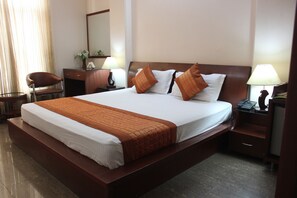 A hotel room with a large bed, a desk with a chair, a nightstand with a lamp, and a mirror on the wall. - Hotel Gokul Residency (New Delhi)