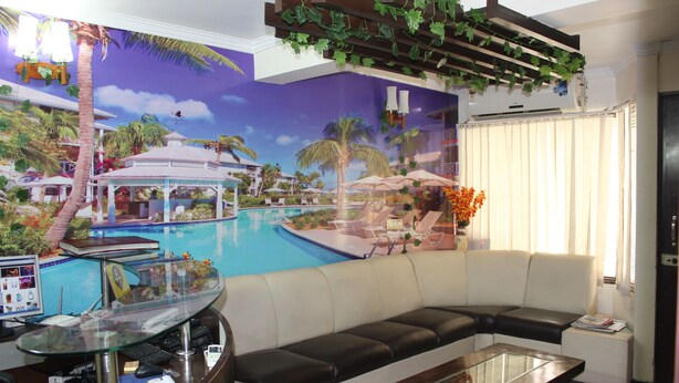 A living room with a curved glass desk, a sofa, and a view of a pool area with palm trees and lounge chairs. - Hotel Gokul Residency (New Delhi)
