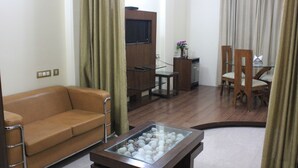 A living room with a brown leather sofa, a glass coffee table with decorative items, a wooden TV stand, and a dining area with a wooden table and chairs. - Hotel Gokul Residency (New Delhi)