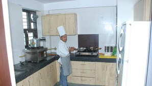 A chef in a kitchen with wooden cabinets, a stove, and a refrigerator. - Hotel Gokul Residency (New Delhi)