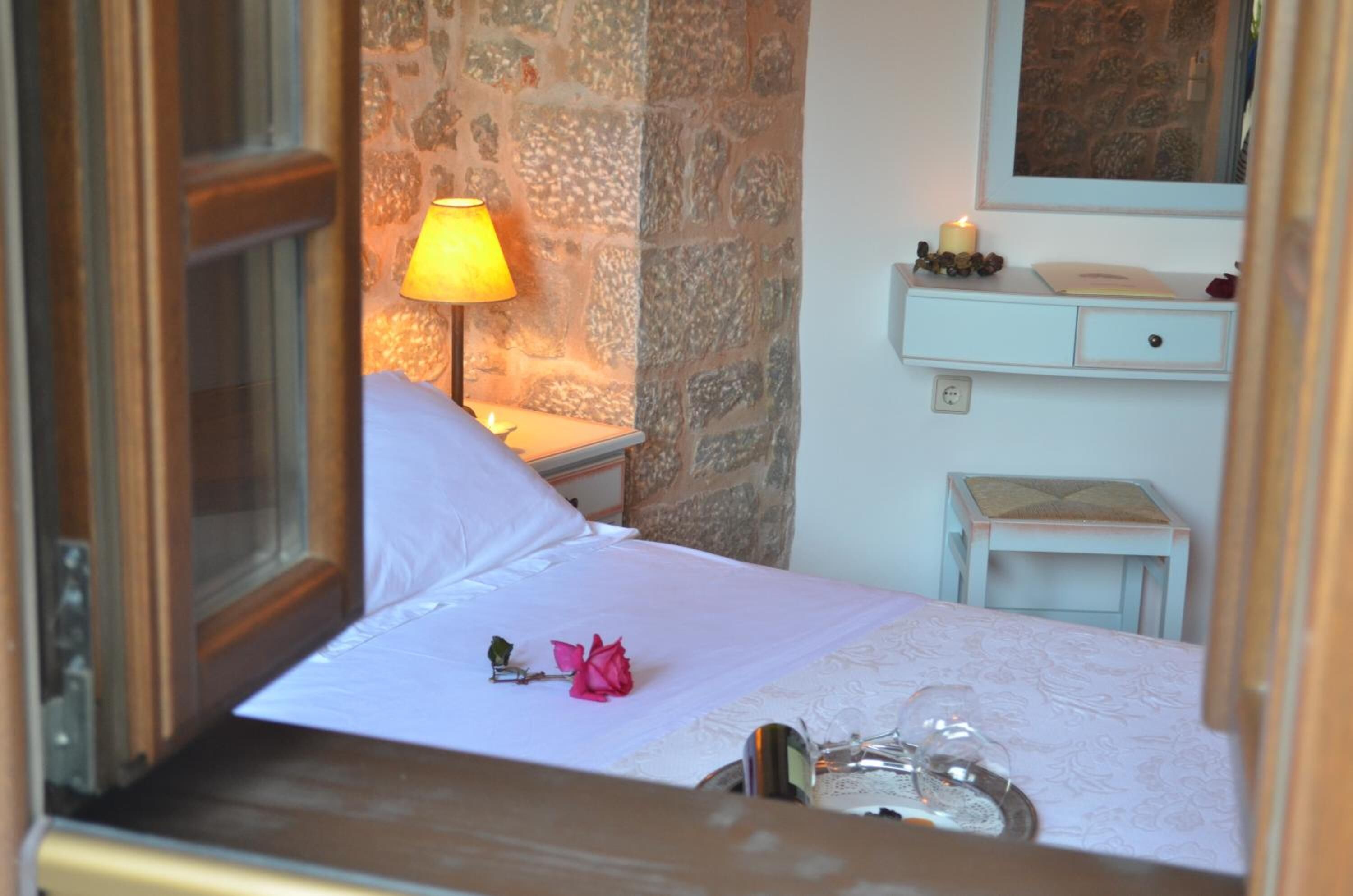 A bedroom with a stone wall, a bedside lamp, a bedside table with a candle, a small stool, and a mirror above the table.