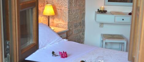 A bedroom with a stone wall, a bedside lamp, a bedside table with a candle, a small stool, and a mirror above the table.