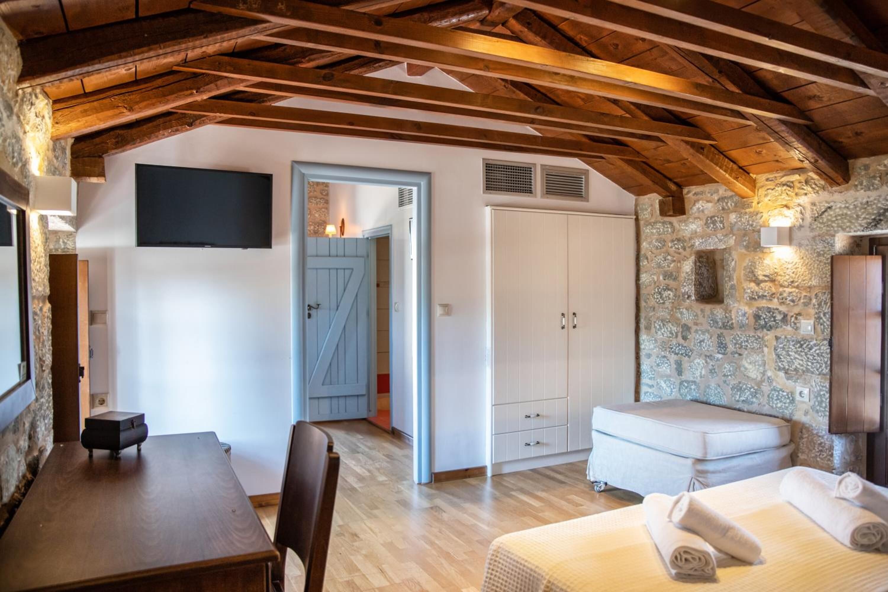A bedroom with a stone wall, wooden ceiling beams, a bed, a small table, and a wardrobe.