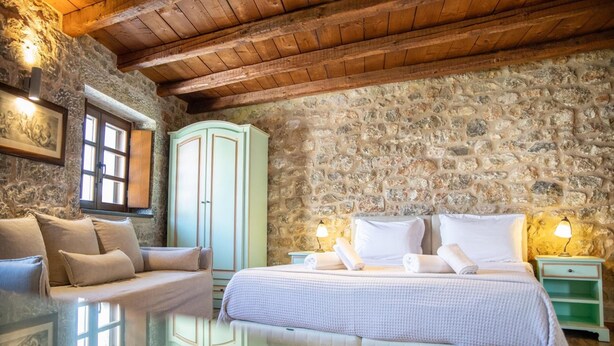 Un dormitorio con pared de piedra, vigas de madera en el techo, una cama con ropa blanca, una mesita de noche verde y una ventana con cortinas.