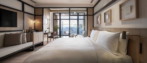 A modern hotel room with a large bed, a desk, and a view of a cityscape through floor-to-ceiling windows.
