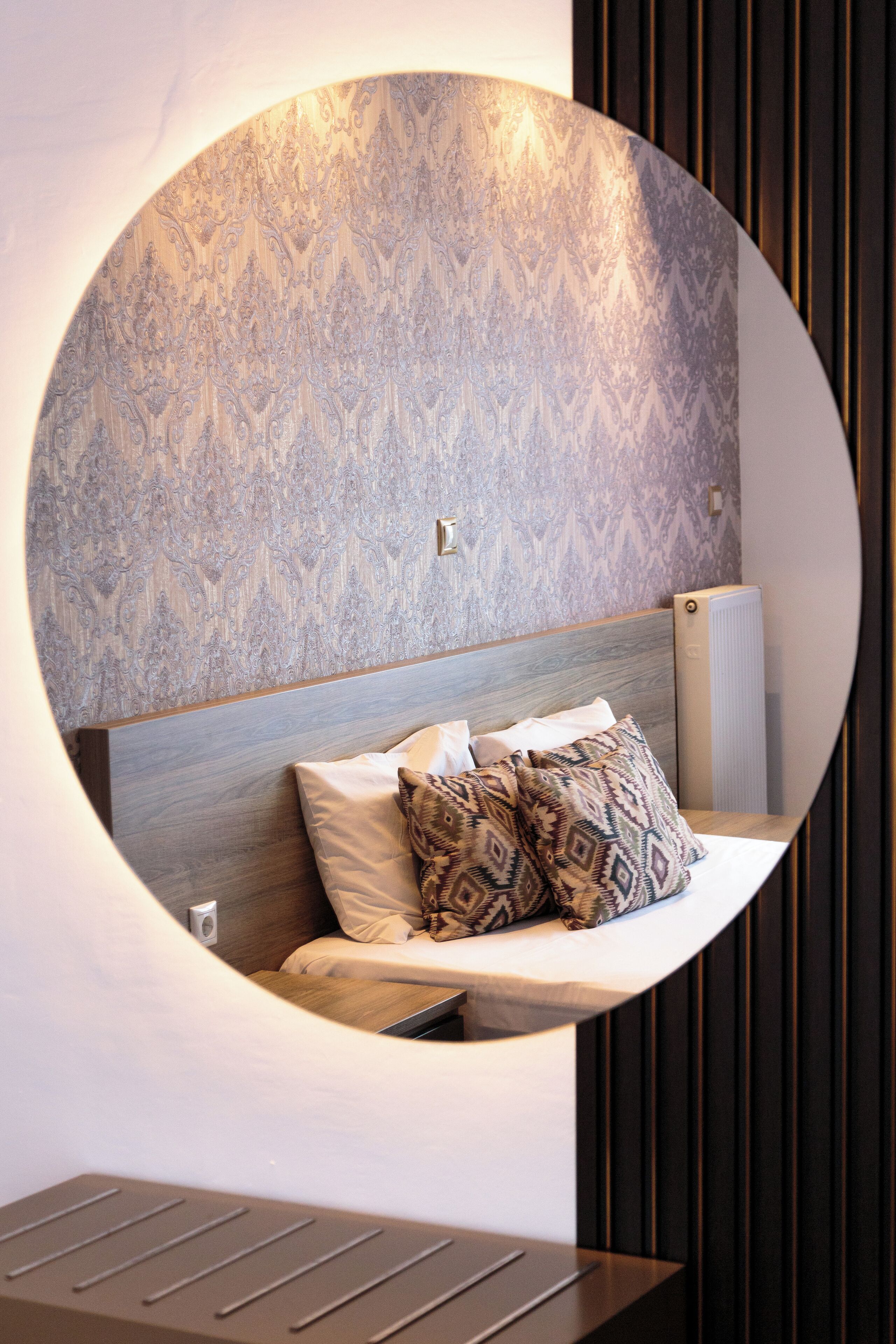 Une fenêtre ronde, un papier peint à motifs, une tête de lit en bois et une table de chevet avec une lampe.