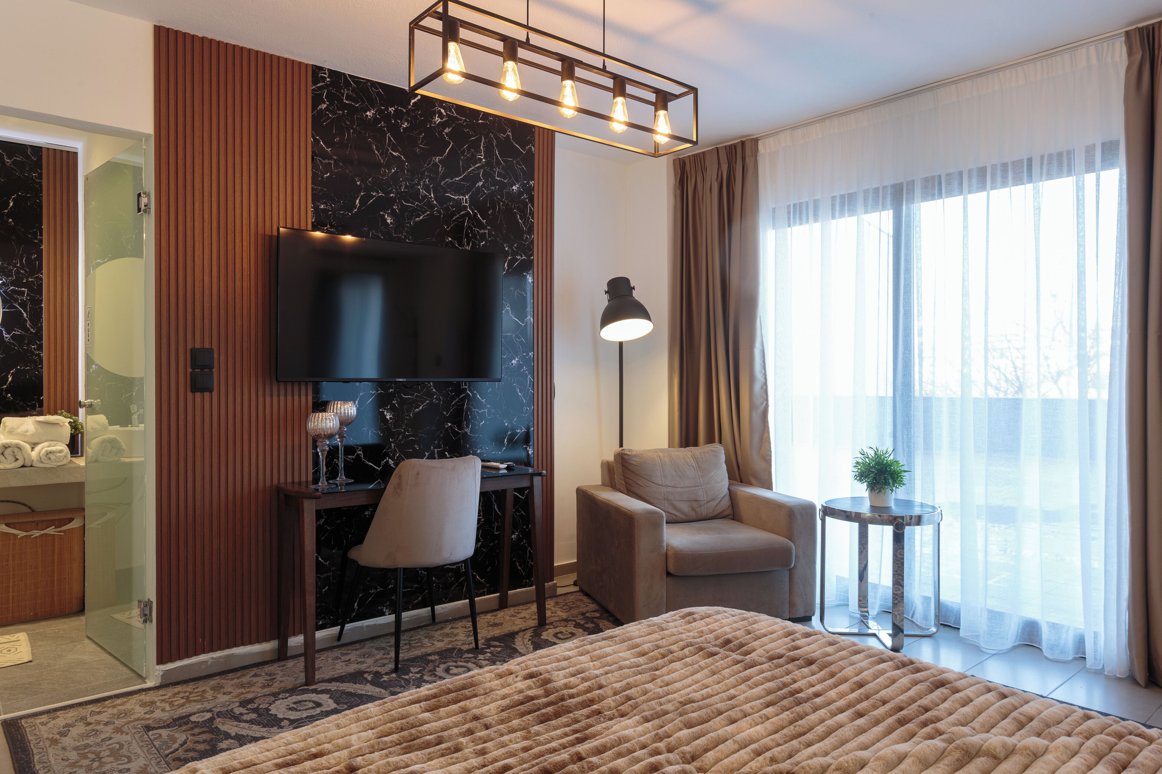 A modern hotel room with a large bed, a flat-screen TV, a desk with a chair, a small side table, and a view of the outdoors through sheer curtains.