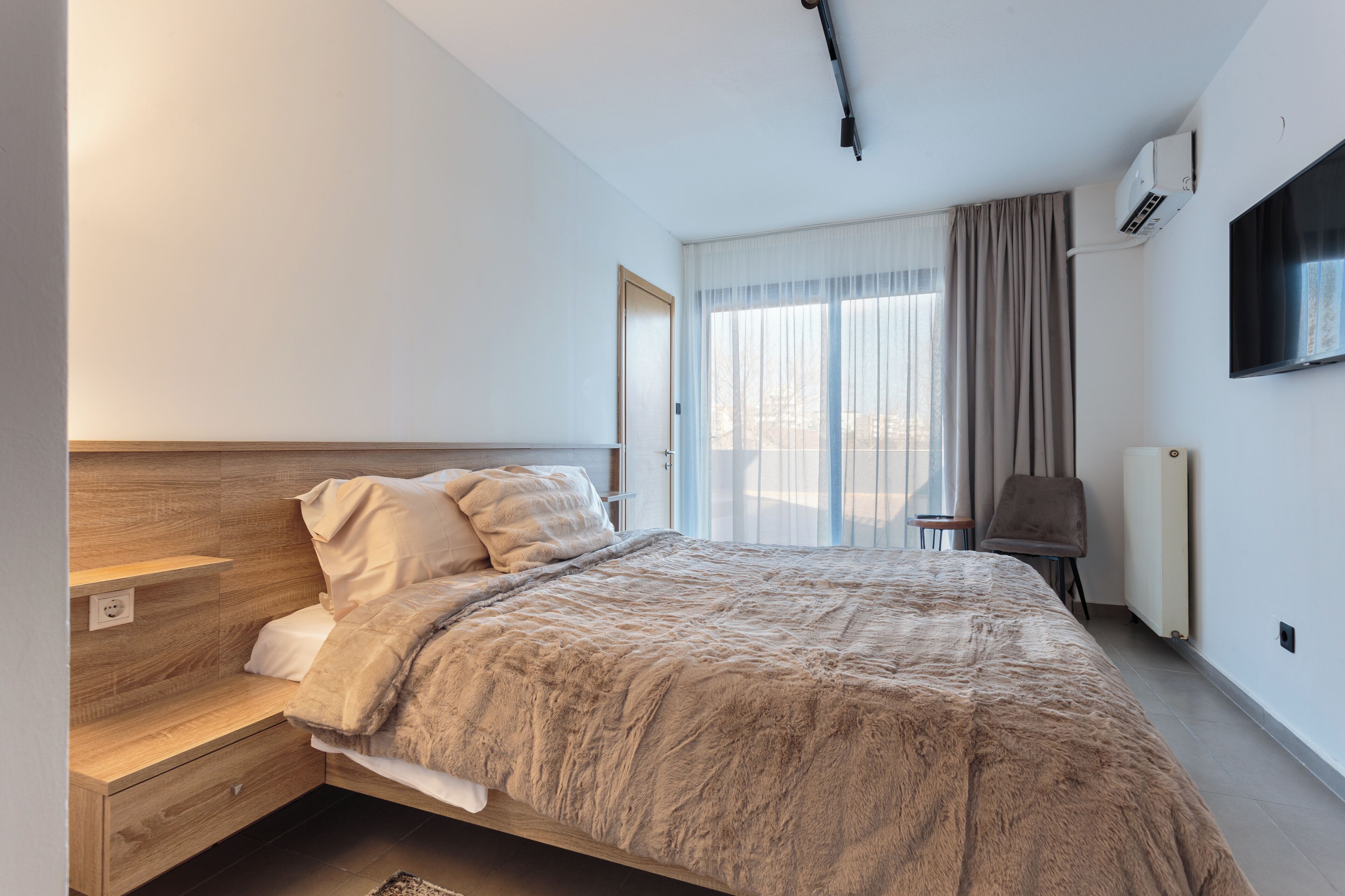 Un dormitorio moderno con cabecera de madera, una cama grande con manta mullida, una mesita de noche, una silla, un televisor montado en la pared y una ventana con cortinas.