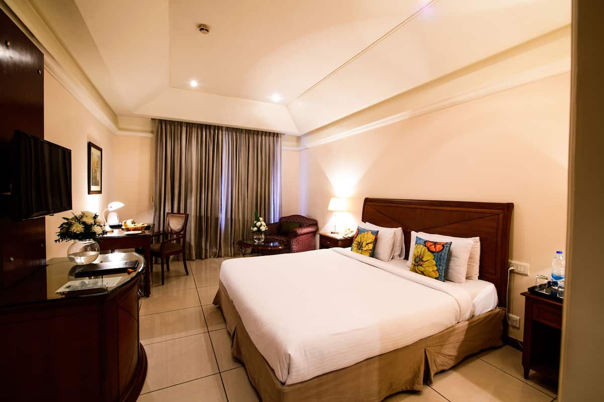 Quarto de hotel com uma cama grande, uma escrivaninha com luminária, uma cadeira, uma televisão e uma janela com cortinas.
