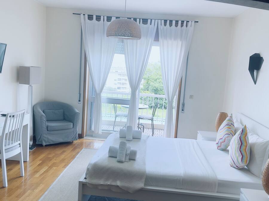 A hotel room with a bed, a desk, a chair, a TV, and a balcony with curtains.
