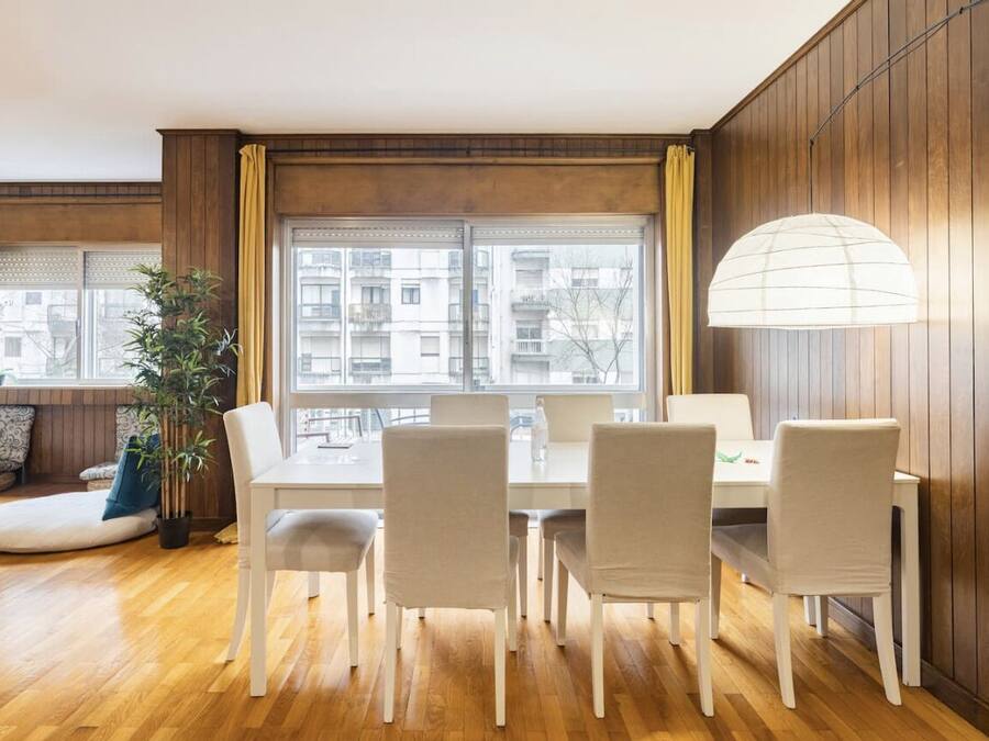 Un comedor con piso de madera, una mesa blanca y cuatro sillas. Se observa un ventanal con cortinas, una planta en maceta y una lámpara colgante.