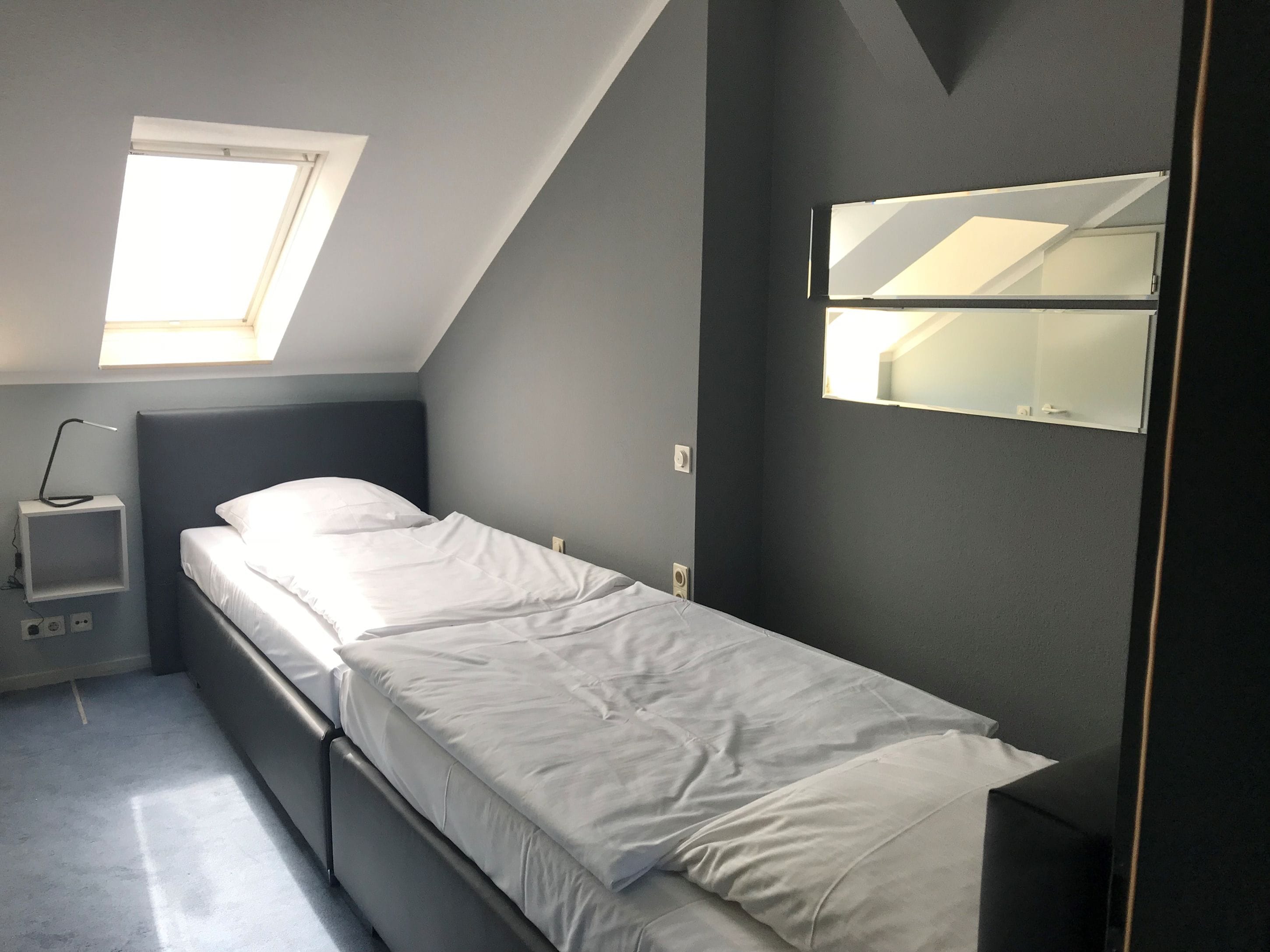 Quarto pequeno com uma cama de solteiro, claraboia, mesa de cabeceira e espelho fixado na parede.