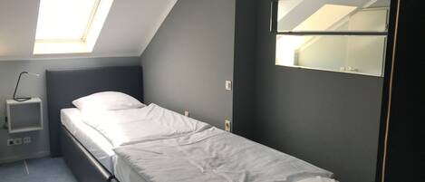 Quarto pequeno com uma cama de solteiro, claraboia, mesa de cabeceira e espelho fixado na parede.