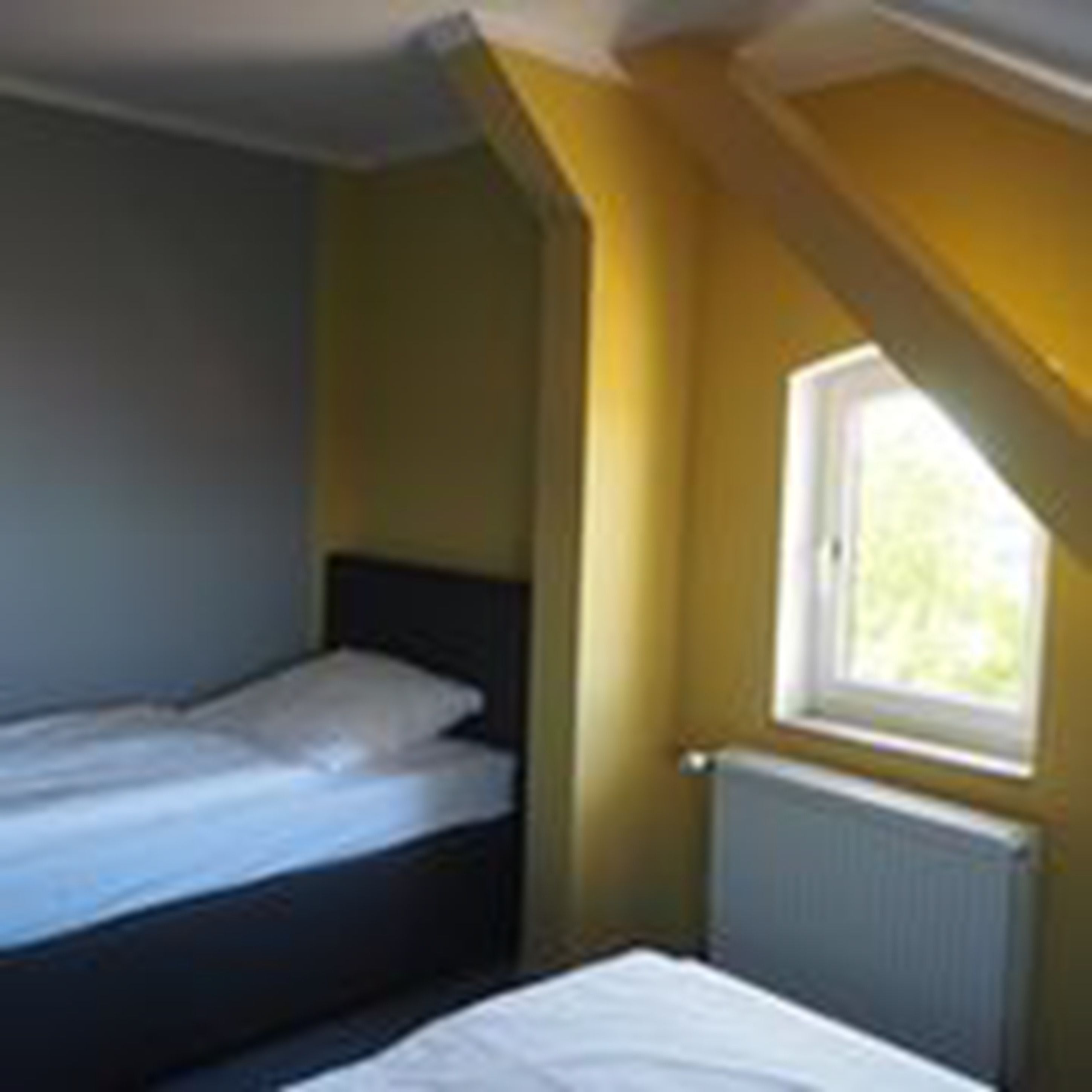 Ein kleines Zimmer mit einem Einzelbett, einem Fenster, einem Heizkörper und einem teilweisen Blick auf eine Tür.