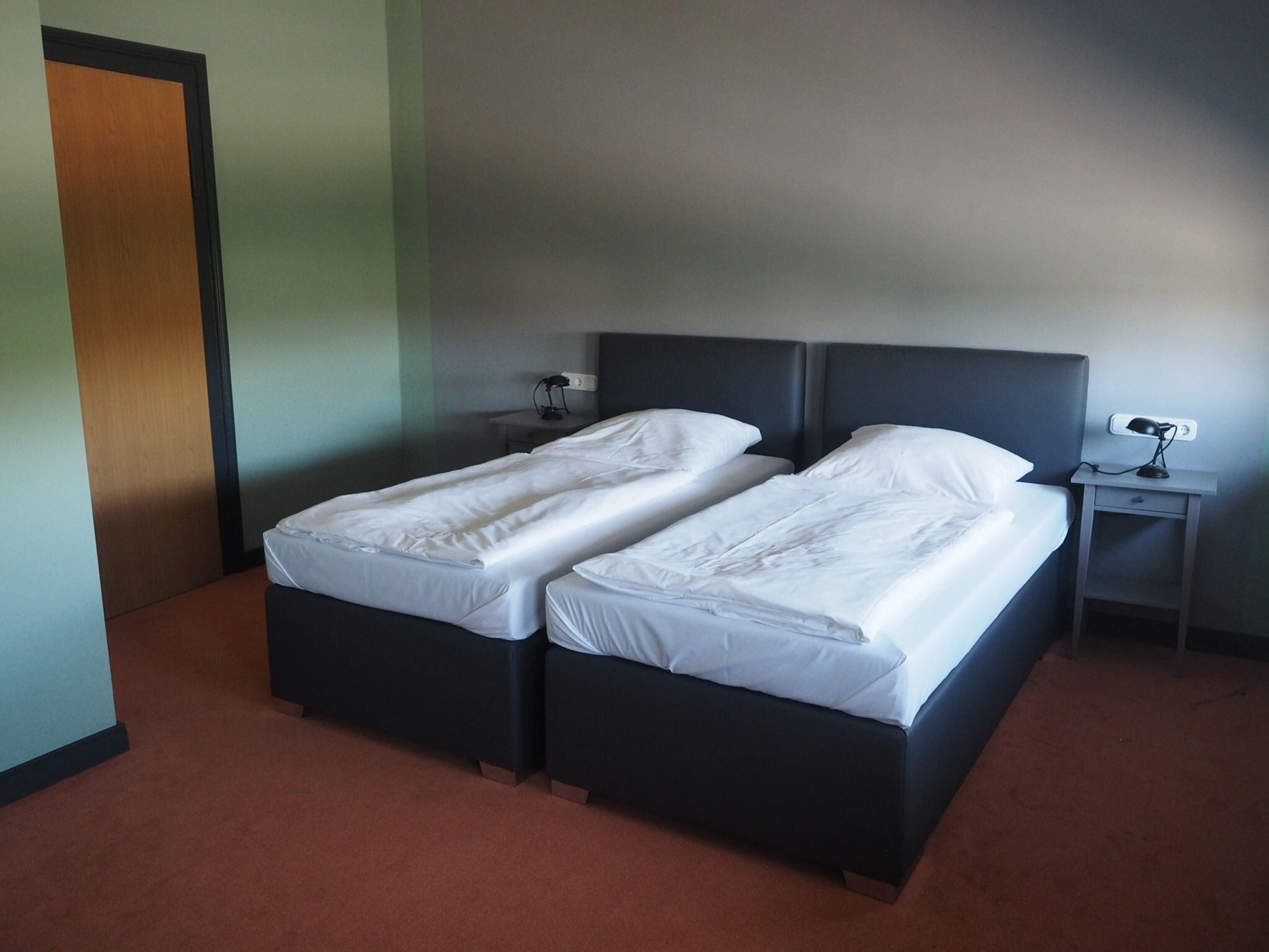 Una habitación con dos camas individuales, una mesita de noche con lámpara y una puerta.