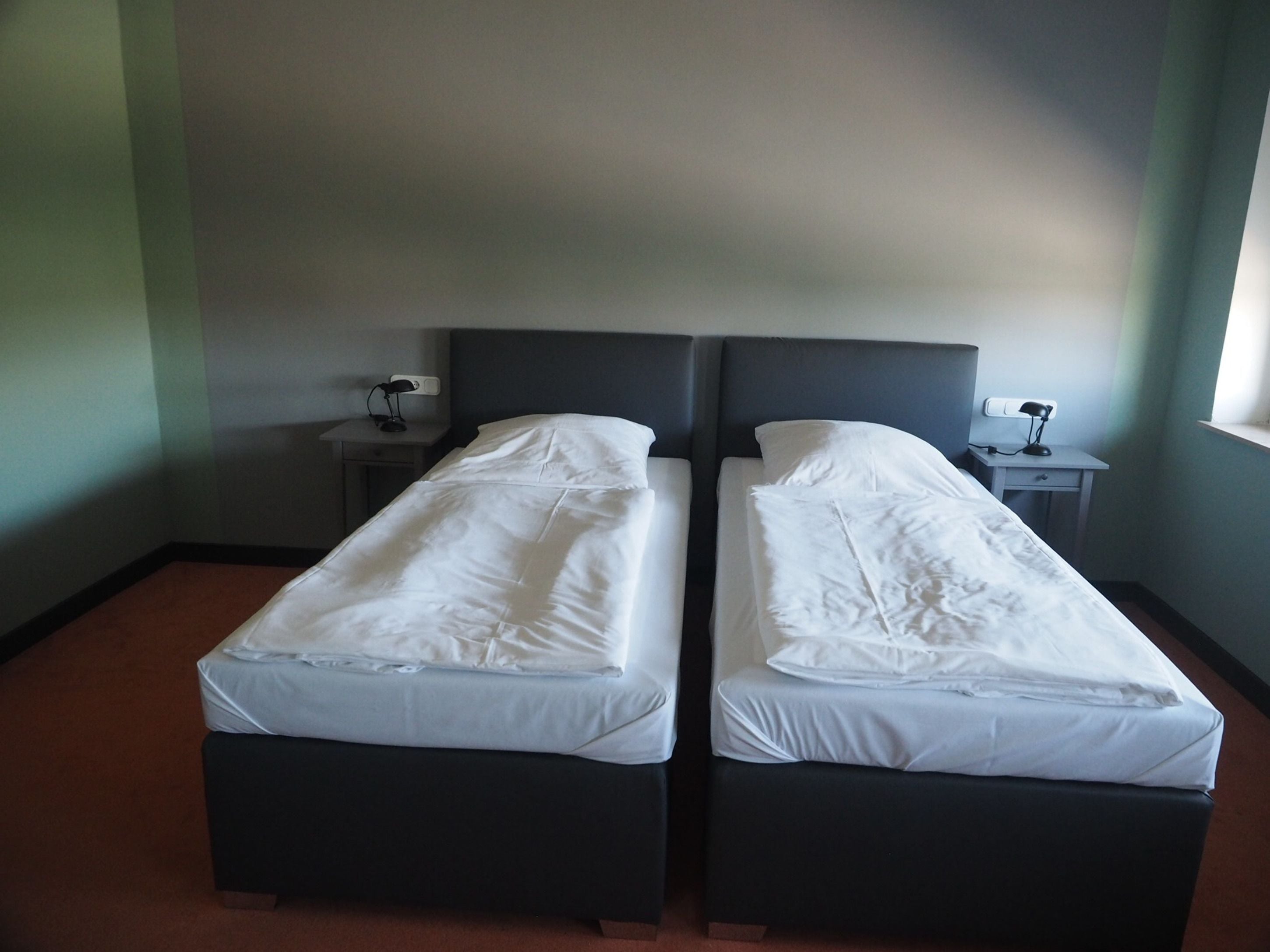 A room with two beds, each with a dark headboard and white bedding. There are two bedside tables with lamps, and a window on the right side.