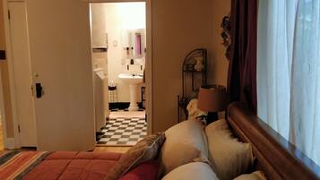 A hotel room with a checkered floor, a bed with red and white bedding, a window with curtains, and a view into the bathroom with a sink and mirror.