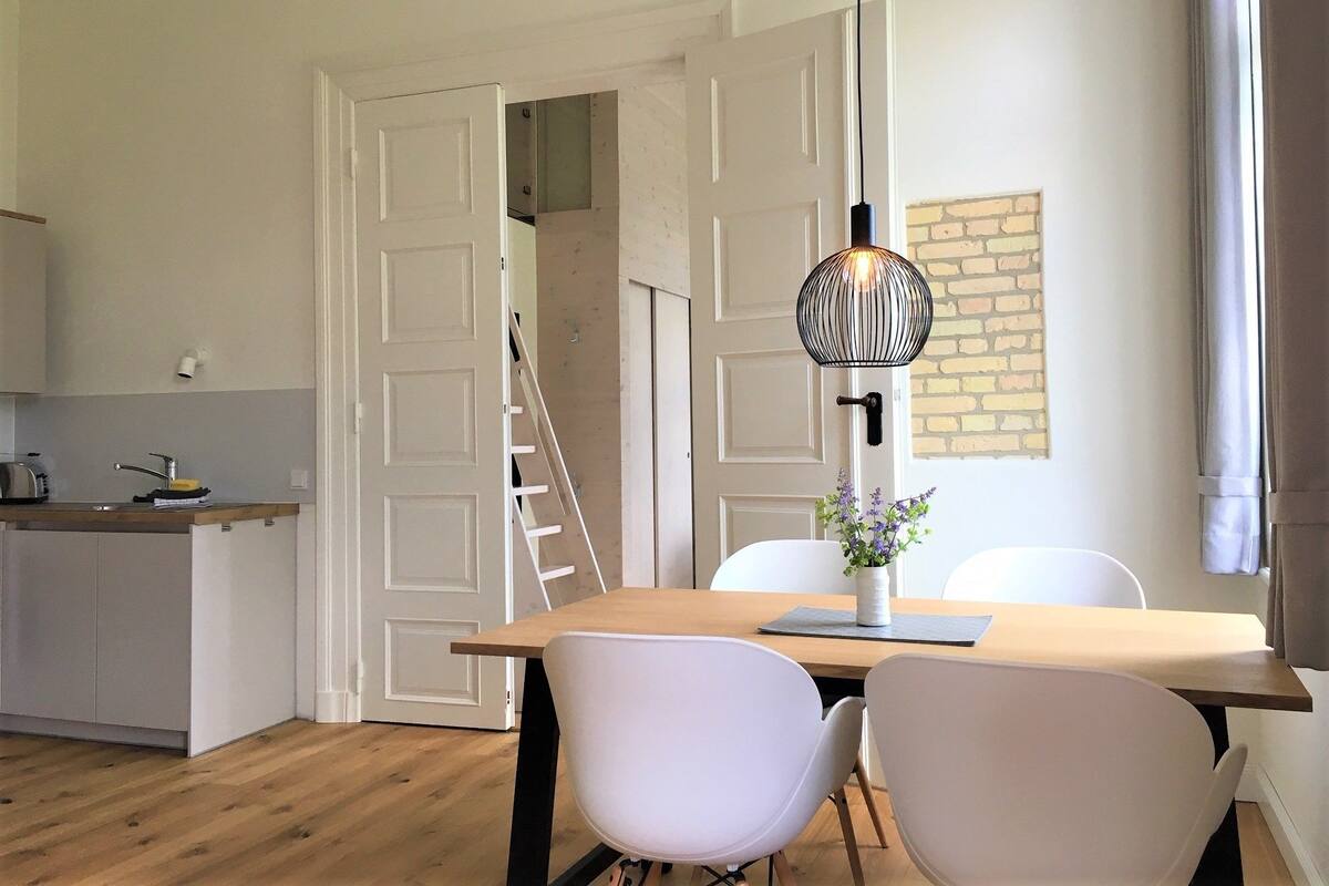 A modern kitchen with white cabinetry, a wooden countertop, and a built-in stove. There is a dining area with a wooden table and two white chairs. A black pendant light hangs above the table.