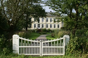 A gated entrance to a large, two-story building with a symmetrical facade, surrounded by lush greenery and trees. - Ferienhof Gut Oestergaard (Steinberg)