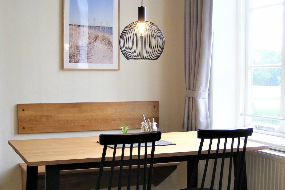 A dining area with a wooden table, two black chairs, a hanging pendant light, and a framed picture on the wall.