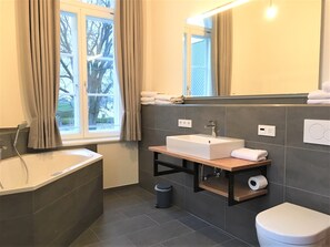 Een moderne badkamer met een badkuip, een wastafel en een toilet. Er is een raam met gordijnen, een spiegel en een handdoekhouder.