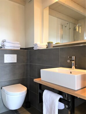 A modern bathroom with a white sink, dark tiled walls, a toilet, and a mirror above the sink. - Ferienhof Gut Oestergaard (Steinberg)