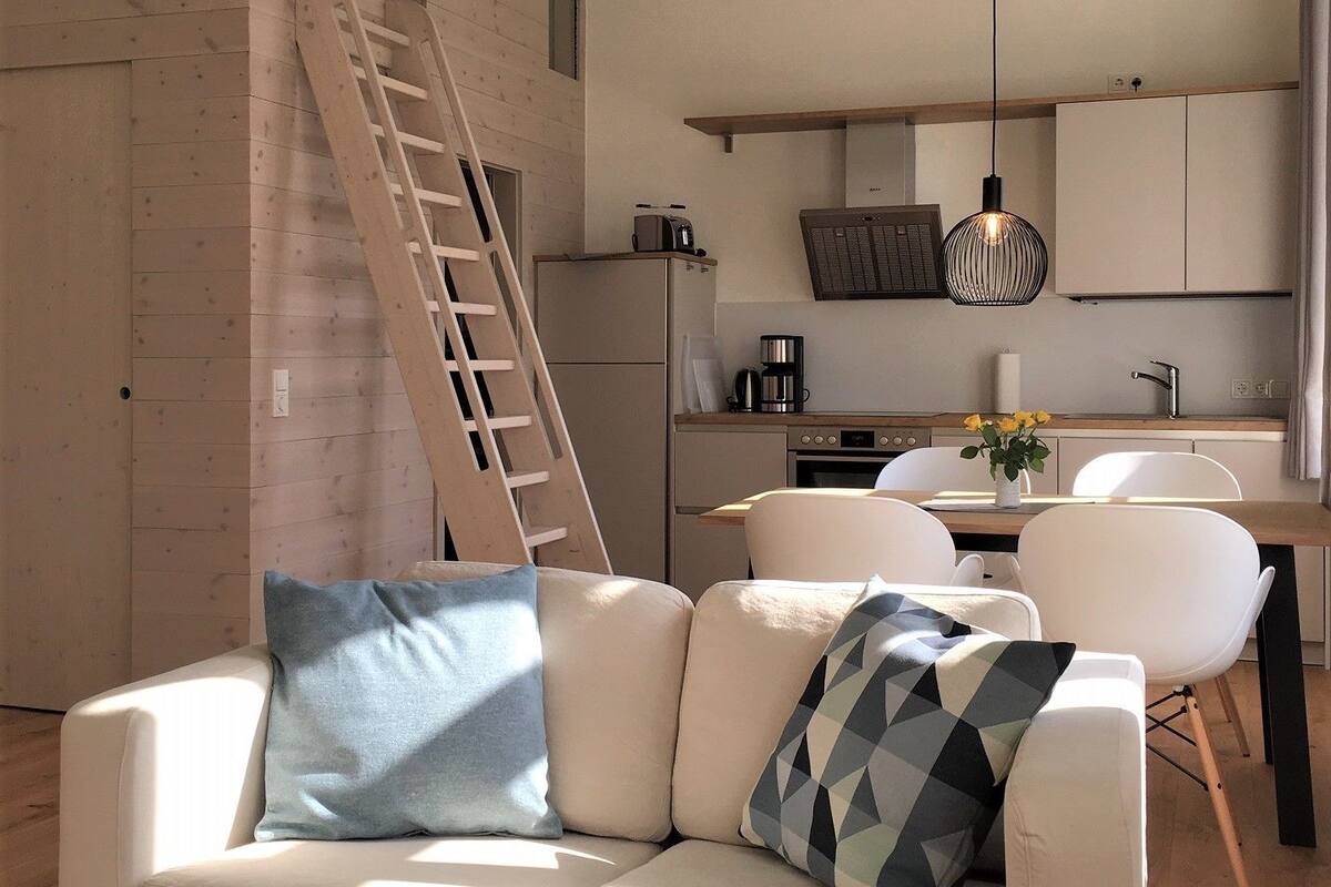 A modern living room with a white sofa, a wooden coffee table, and a kitchen area with a ladder leading to an upper level.