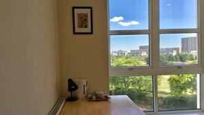 A room with a wooden desk, a radiator, a framed picture, and a window with a view of buildings and trees. - Nice and private rooms (London)