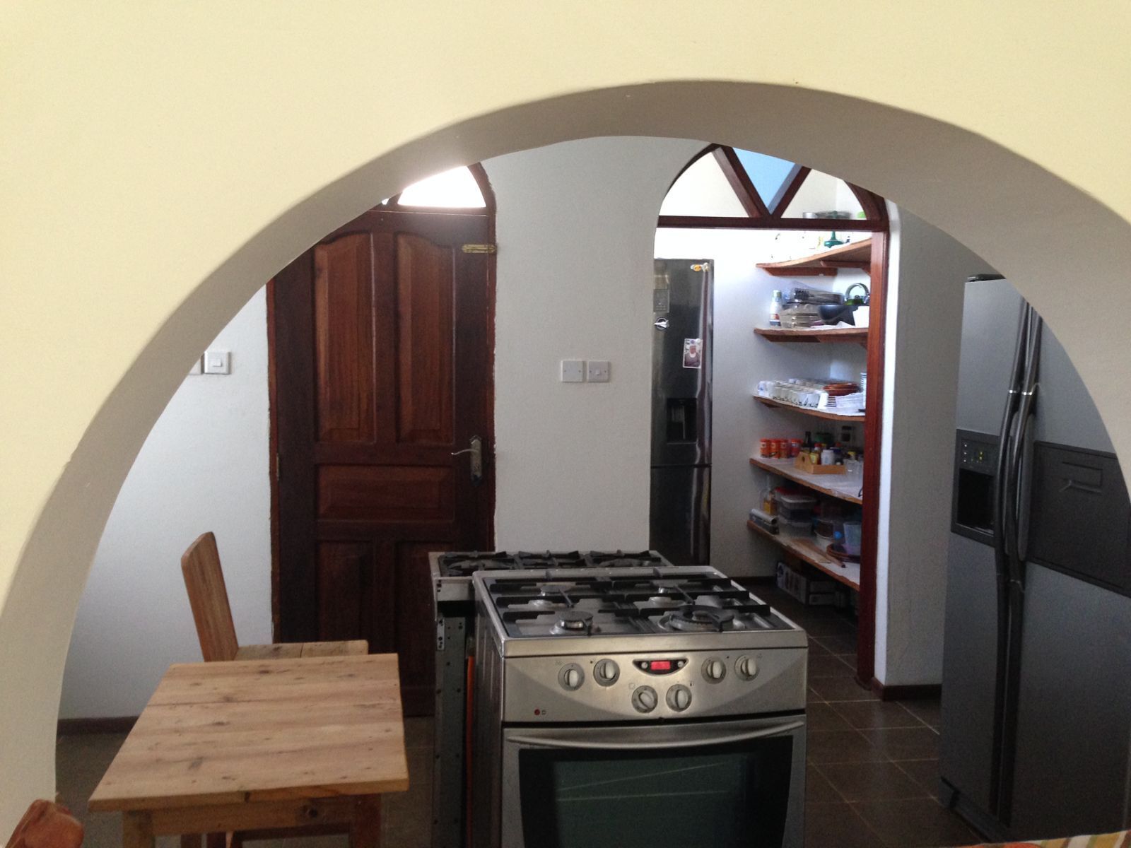 A compact kitchen with a gas stove, oven, and a refrigerator. There is a wooden table and a chair, and a wooden door leading to another room.