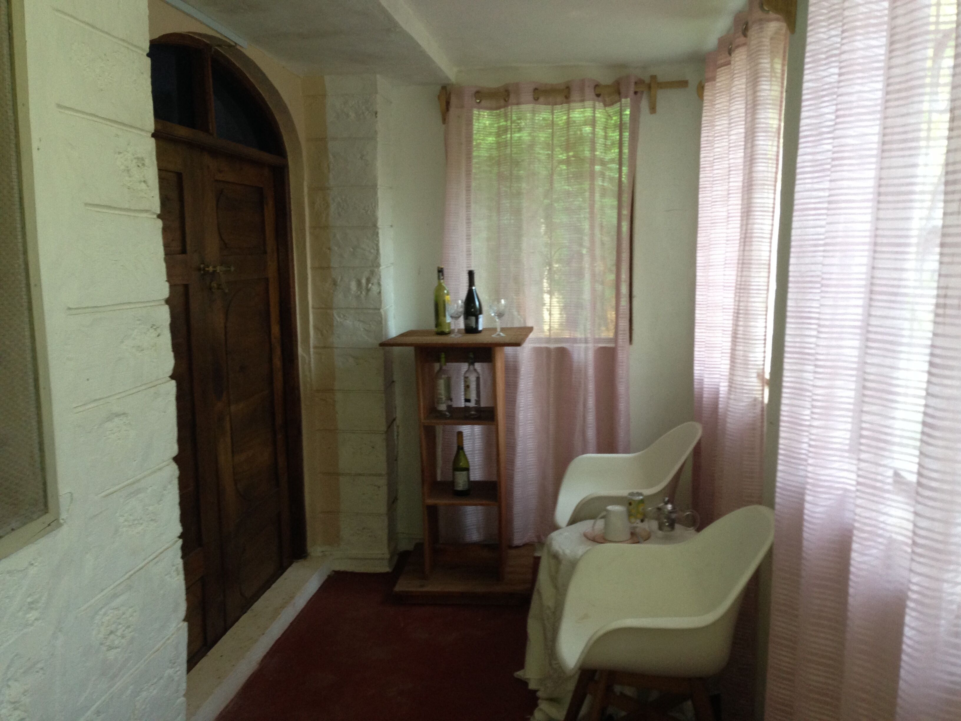 A narrow room with a wooden door, a small table with wine bottles and glasses, and two white chairs.