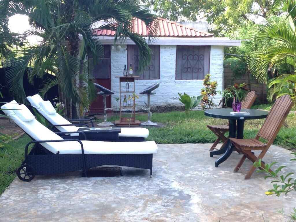 Um pátio com duas poltronas, uma mesa e uma cadeira. Há um pequeno jardim com uma palmeira e uma casa ao fundo.