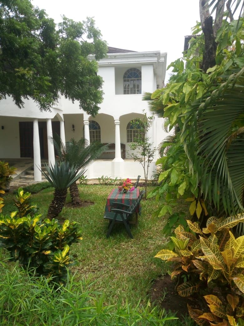A courtyard with a covered seating area, surrounded by lush greenery and a white building with arched windows.